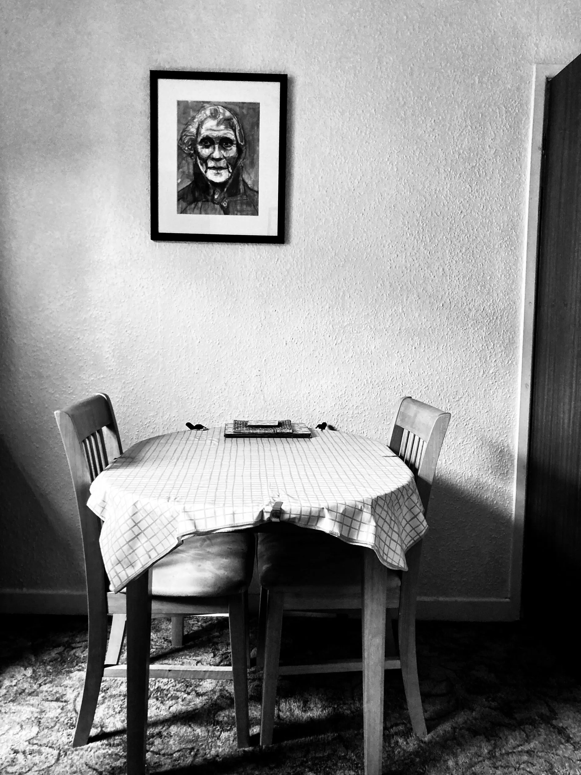 A black and white photograph of a round dining table with two chairs with a portrait hanging on the wall above.
